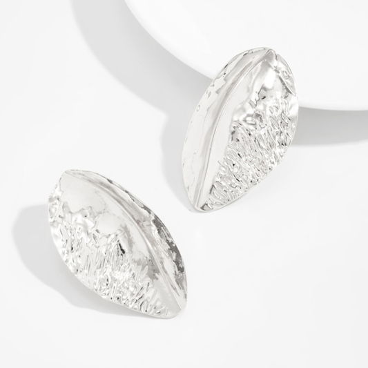 Alba silver-tone sculptural earrings with curved, leaf-like shapes and hammered texture, displayed beside seashells on a cream background