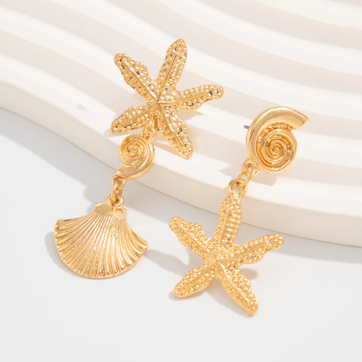 Estrella asymmetrical gold-tone drop earrings with textured starfish, spiral and scallop shell charms on a white surface.