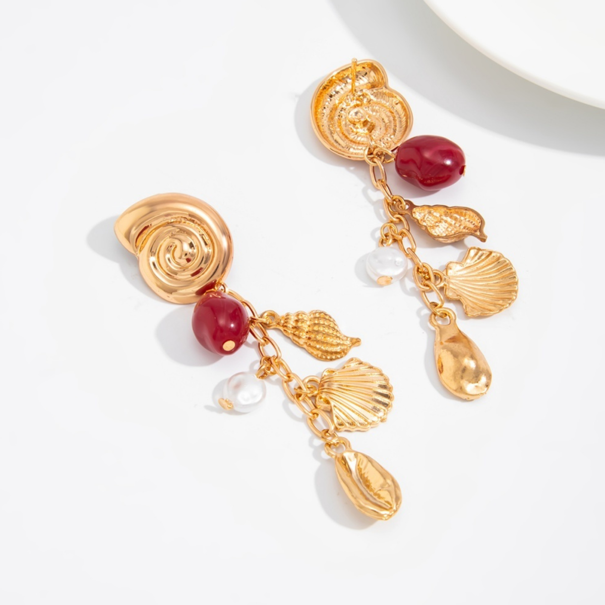 Marina gold-tone drop earrings with spiral studs, shell charms, deep red beads and pearl-look accents, shown on a model