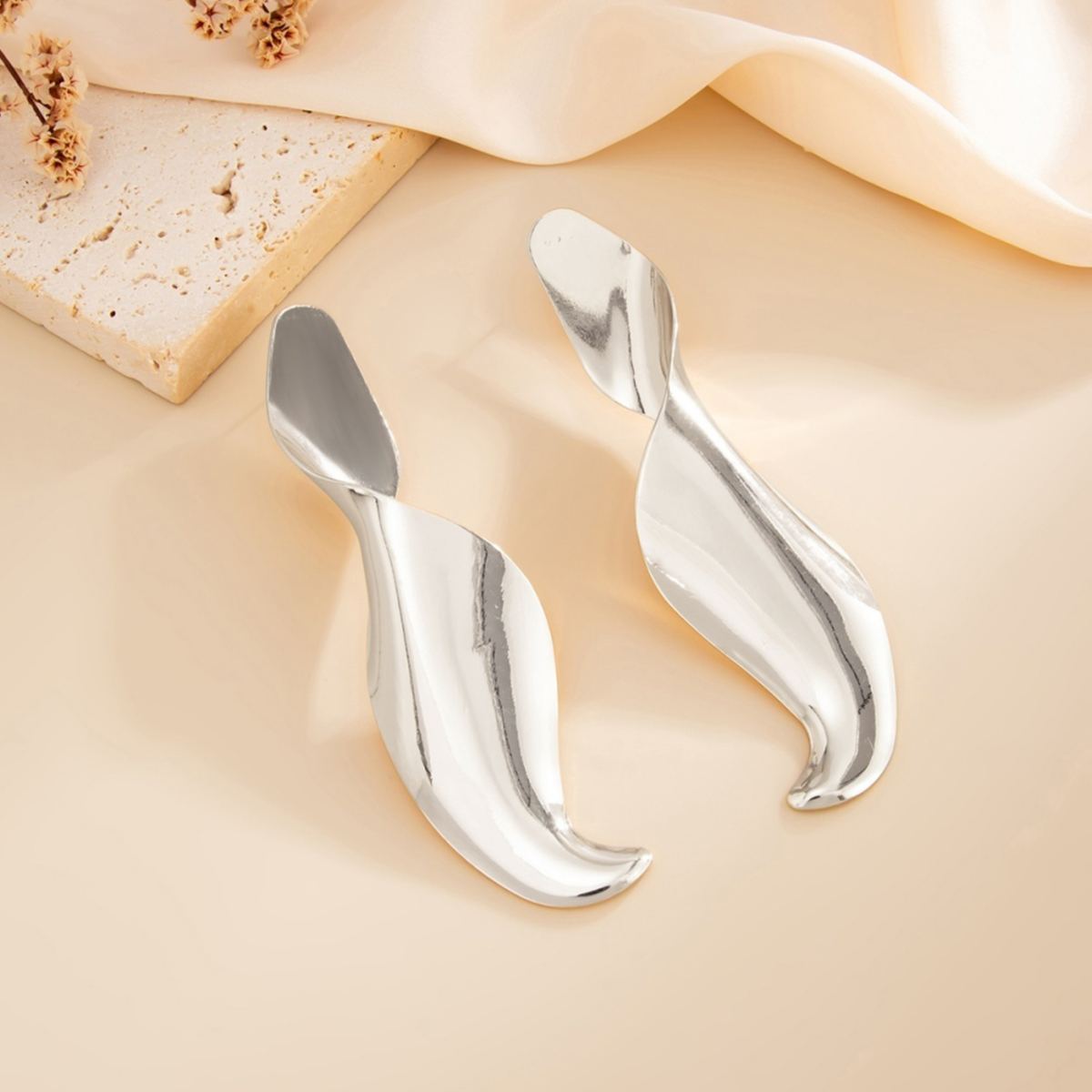 Pair of Soleil silver-tone sculptural drop earrings with a polished, twisted ribbon shape, arranged on a cream background.